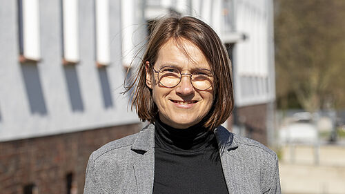 Es ist die Professorin zu sehen, die vor einem Gebäude steht. Ihre Haare werden vom Wind leicht über das Gesicht geweht.