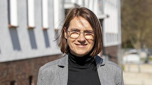 Es ist die Professorin zu sehen, die vor einem Gebäude steht. Ihre Haare werden vom Wind leicht über das Gesicht geweht.