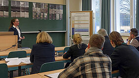 Sandra Jäntsch steht vor einer Tafel und schaut ins Publikum. Hinter ihr sind Bilder von Betonproben sowie ein Poster zur Methodik zu erkennen.