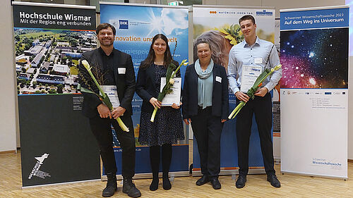 Alle genanntenPErsoenen stehen nebeneinander. Die drei Ausgezeichneten halten Blumen und Urkunden in der Hand. Hinter ihnen stehen vier unterschiedlich gestaltete Rollups.