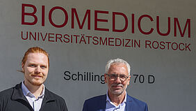 Die beiden Personen sind Porträt nebeneinander zu sehen. Auf der Wand hinter ihnen kann man den Schriftzug „Biomedicum“ lesen.