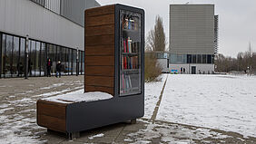 Hinter dem Bücherschrank sieht man links die Glasfassade des Hauses 6 und im Hintergrund das Haus 7. Alles ist mit einer dünnen Schneeschicht bedeckt.