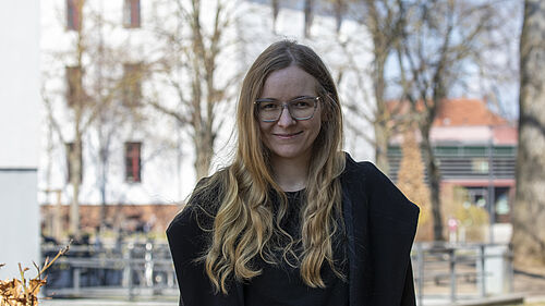Die junge Frau hat einen dunklen Mantel an und steht im Schatten eines Gebäudes. Im Hintergrund sind einige sonnenbeschiene Gebäude der Hochschule zu sehen.