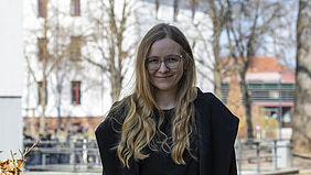 Die junge Frau hat einen dunklen Mantel an und steht im Schatten eines Gebäudes. Im Hintergrund sind einige sonnenbeschiene Gebäude der Hochschule zu sehen.