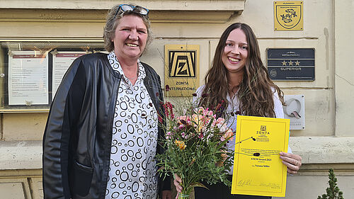 Verena steht gemeinsam mit der ZONTA-Präsidentin vor einer Hauswand und hält die Auszeichnungsurkunde und einen Blumenstrauß in der Hand.