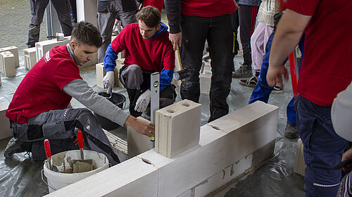 Zwei Studierende hocken hinter einer flachen BEtonwand. Einer hält eine Wasserwaage daran und ein anderer rührt mit einer Kelle in einem Mörteleimer. Weiter stehen um die beiden herum und beobachten sie.